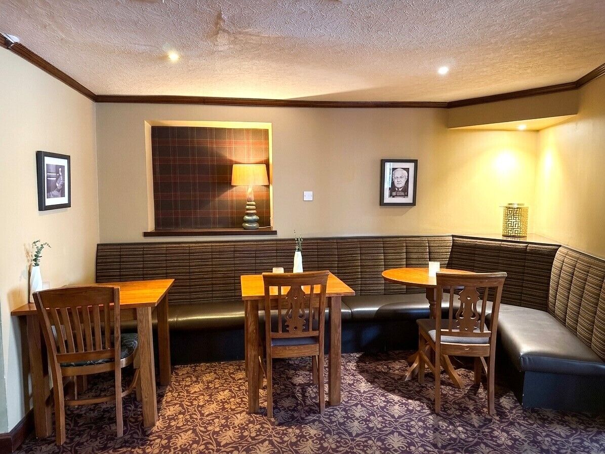 A room with a dark-colored sofa, two wooden tables, and chairs. There are framed pictures on the walls, a lamp on one table, and a patterned carpet.