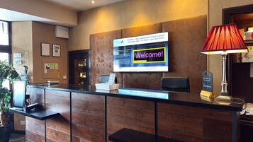 Mostrador de recepción de hotel con una pantalla digital mostrando un mensaje de bienvenida, una lámpara roja y un letrero con el nombre del hotel.