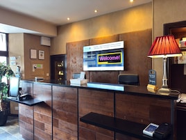Mostrador de recepción de hotel con una pantalla digital mostrando un mensaje de bienvenida, una lámpara roja y un letrero con el nombre del hotel.