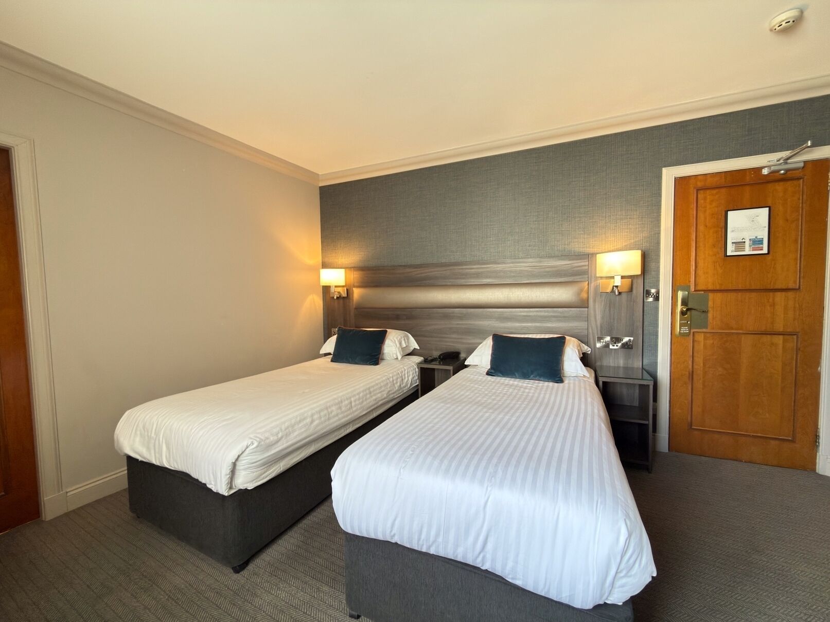 A hotel room with two single beds, a wooden door, and a nightstand with a lamp.