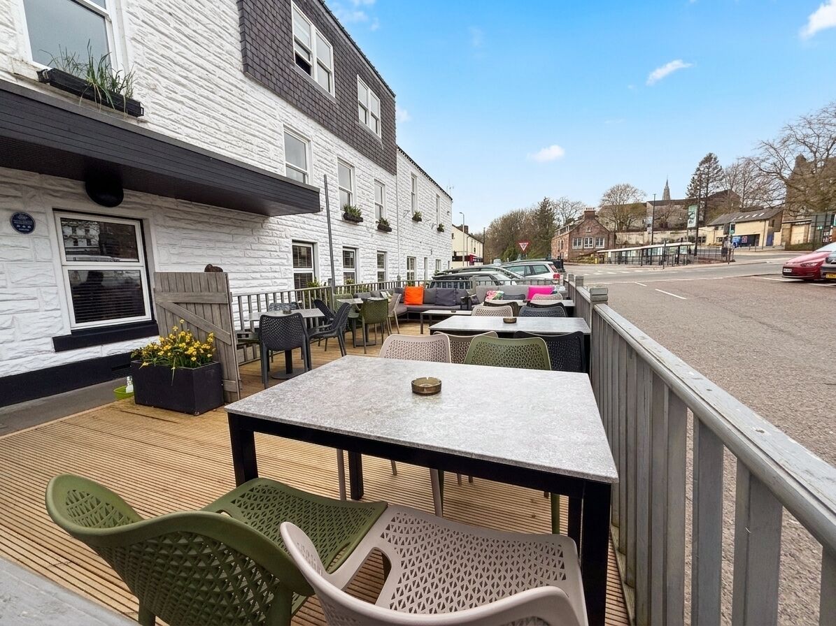 An outdoor seating area with tables and chairs, a building with a white facade, and parked cars in the background.