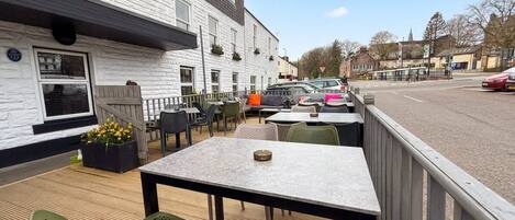 An outdoor seating area with tables and chairs, a building with a white facade, and parked cars in the background.