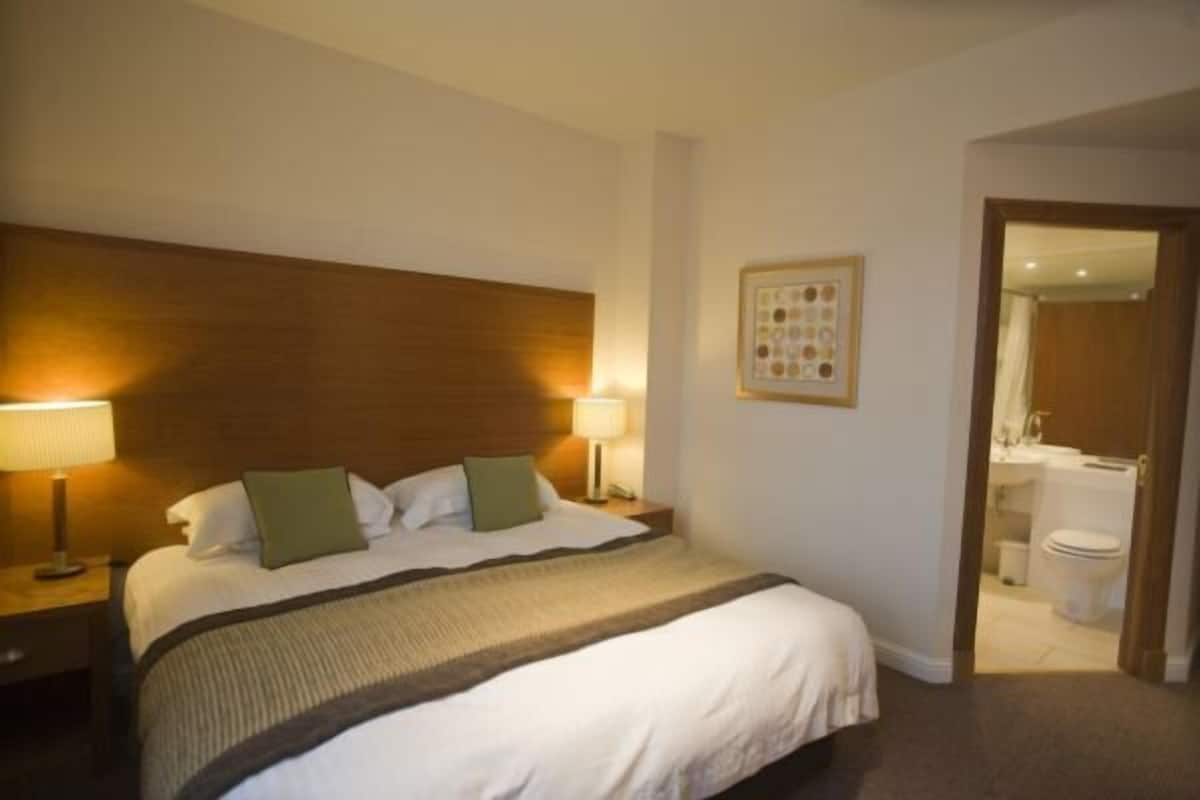 A hotel room with a large bed, two bedside lamps, a wooden headboard, a framed picture on the wall, and a bathroom visible through an open door.