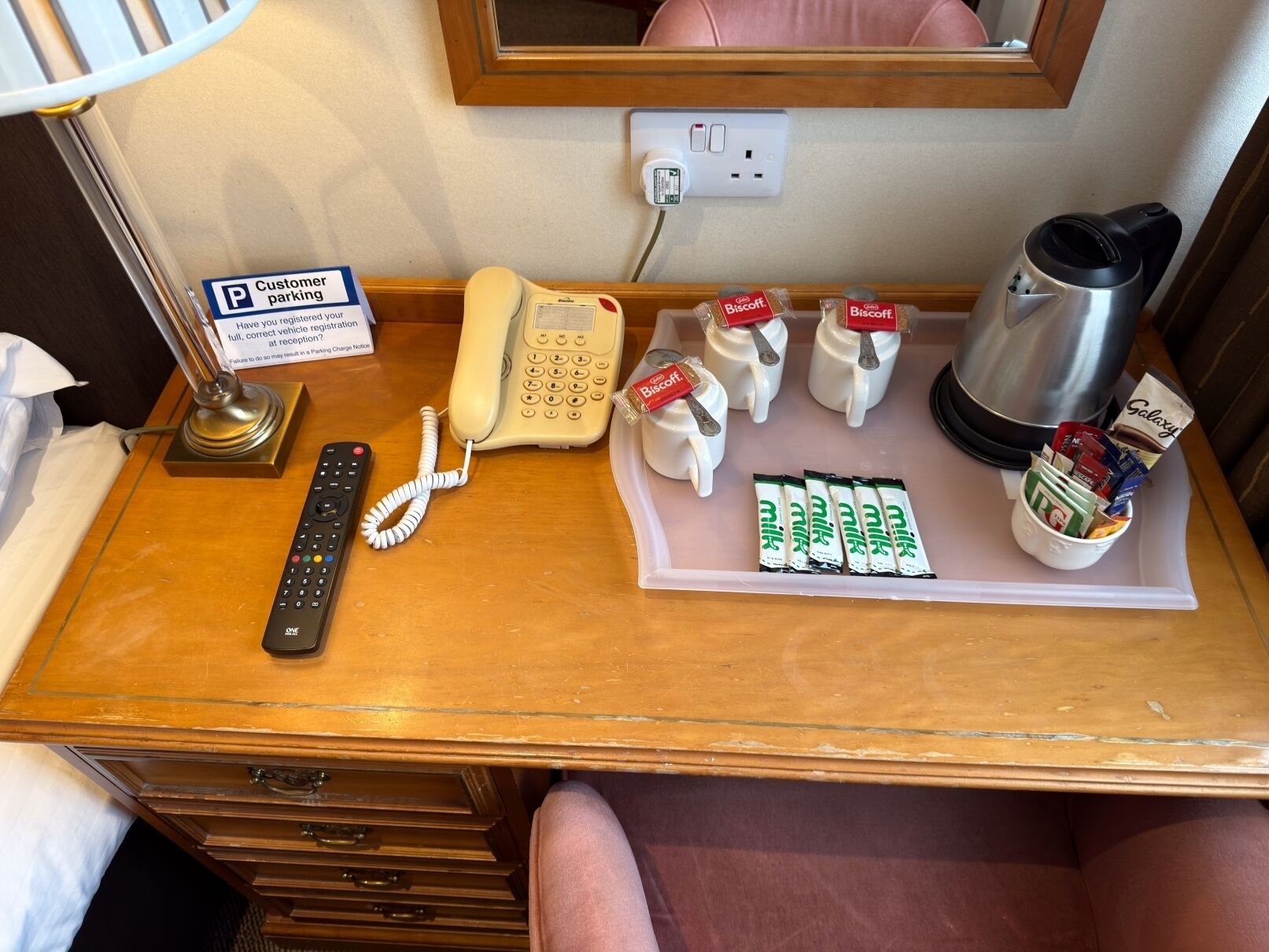 Escritorio de habitación de hotel con teléfono, cartel de estacionamiento para clientes, cafetera y bandeja con leche en sobres y galletas.
