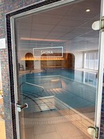 Puerta de cristal que da a un área de sauna con piscina, bancos de madera y un letrero que dice 'SAUNA'.