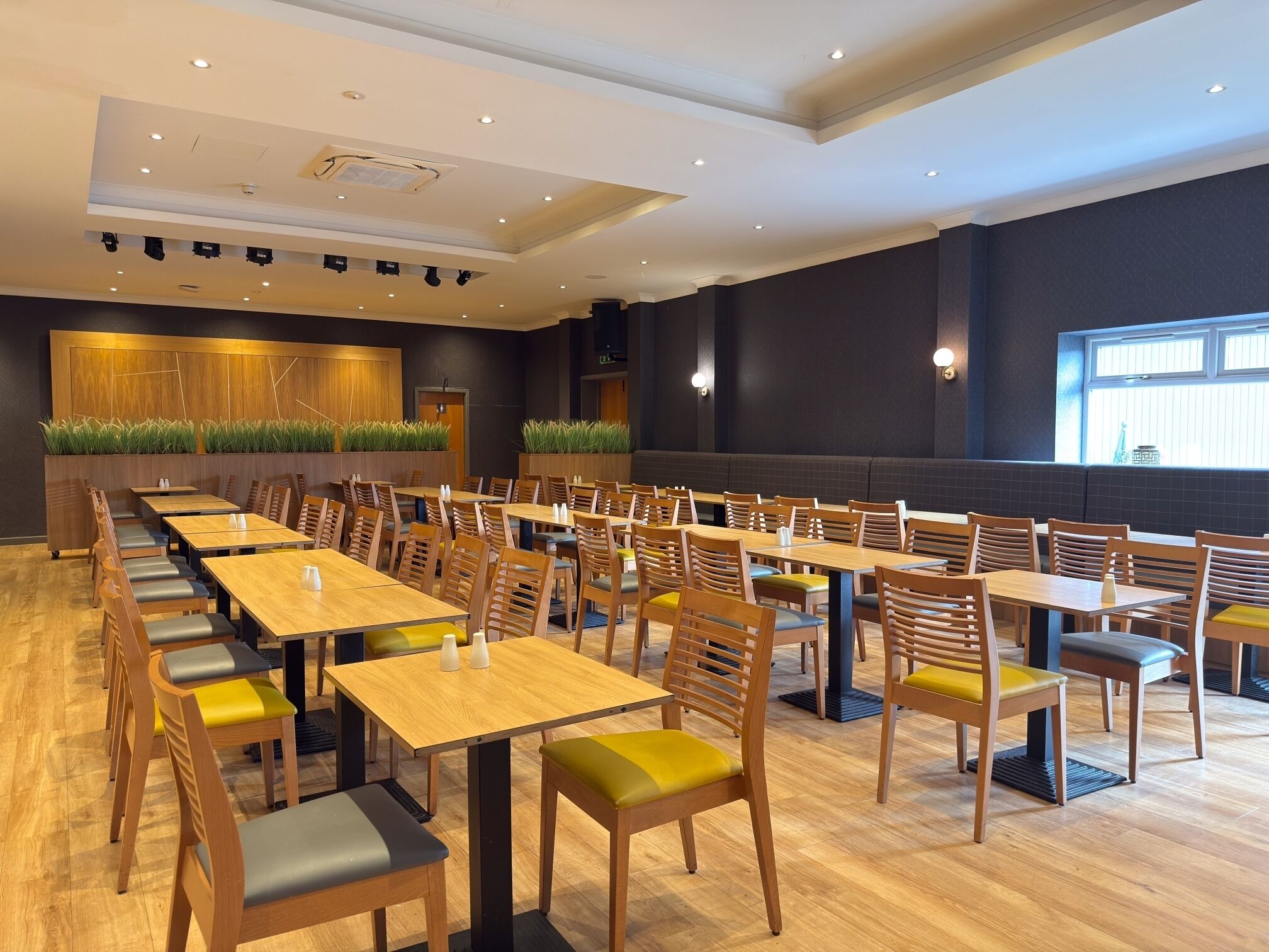 A spacious dining area with multiple tables and chairs, a wooden floor, and a large window providing natural light.