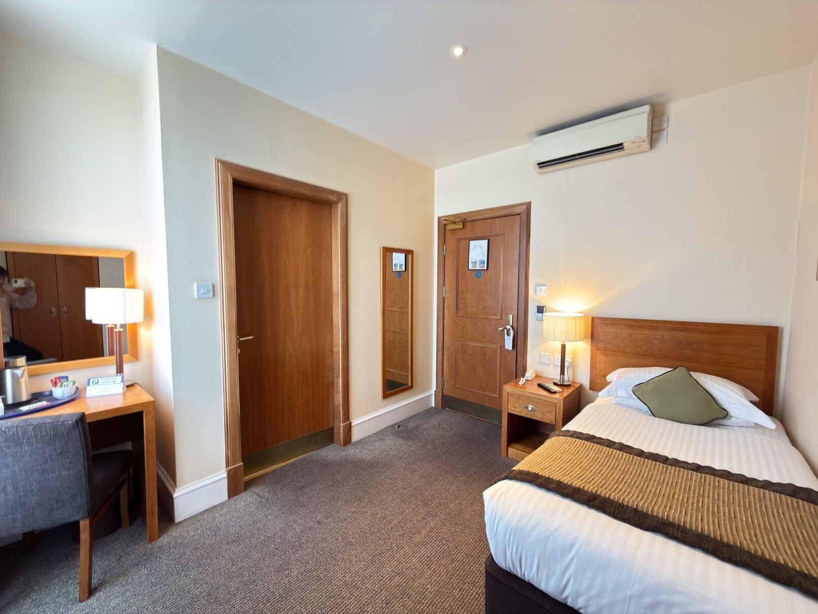 A hotel room with a bed, bedside table, lamp, desk, chair, and a mirror.