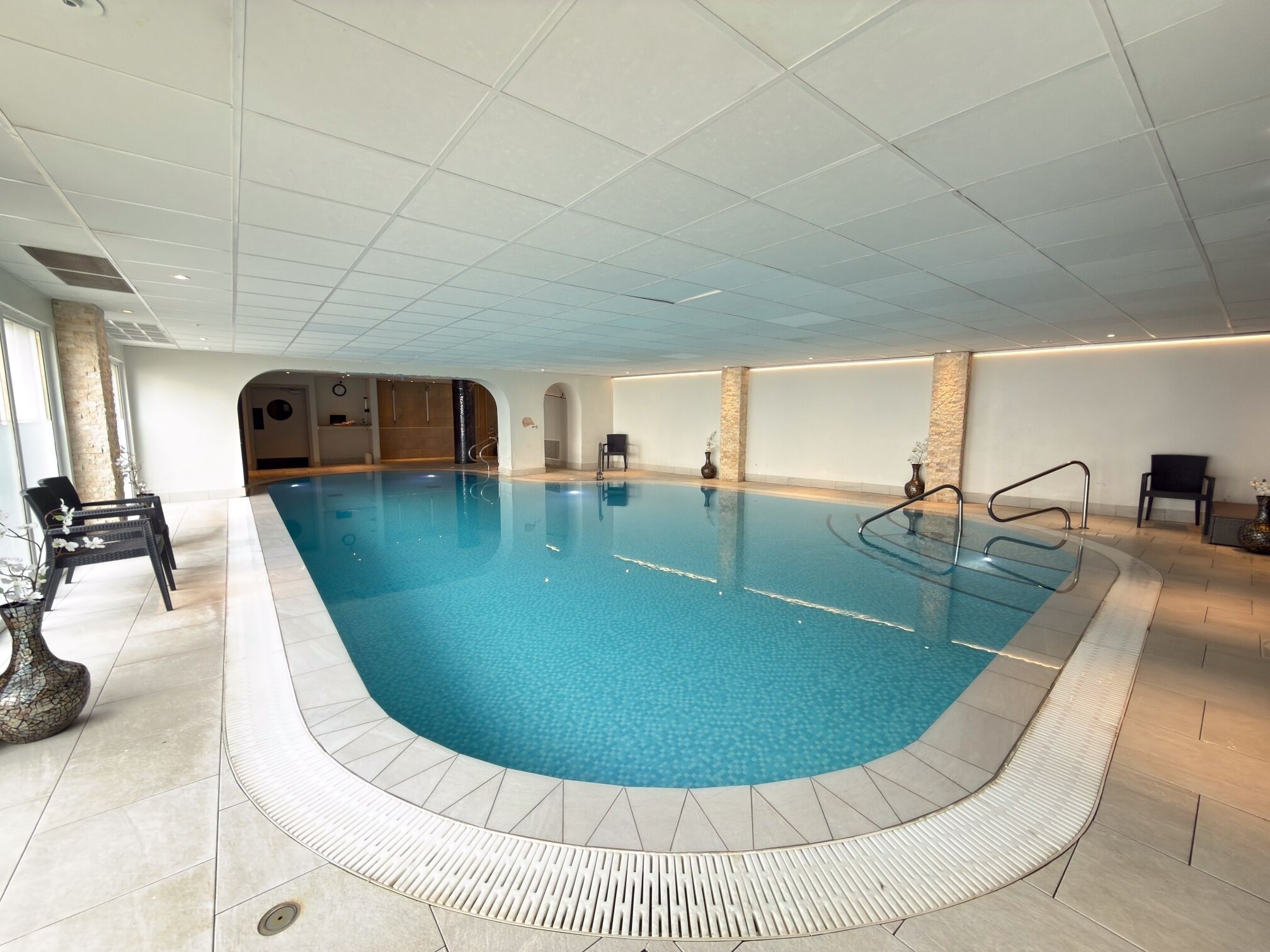 An indoor swimming pool with a tiled floor, a pool ladder, and a few chairs.