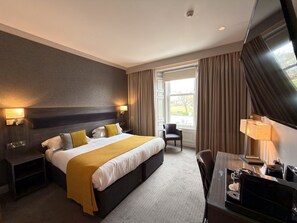 A hotel room with a large bed, bedside tables, a desk with a chair, a TV, and a window with curtains. - The Angus Hotel & Spa by Compass Hospitality (Blairgowrie)