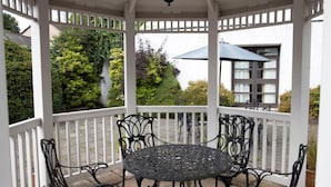 Een overdekt terras met een ronde tafel en stoelen, omgeven door een witte balustrade, met uitzicht op een tuin en een gebouw.