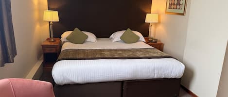 Et hotelværelse med en stor seng, to sengeborde med lamper, en telefon, et billede i ramme på væggen og en lyserød stol.