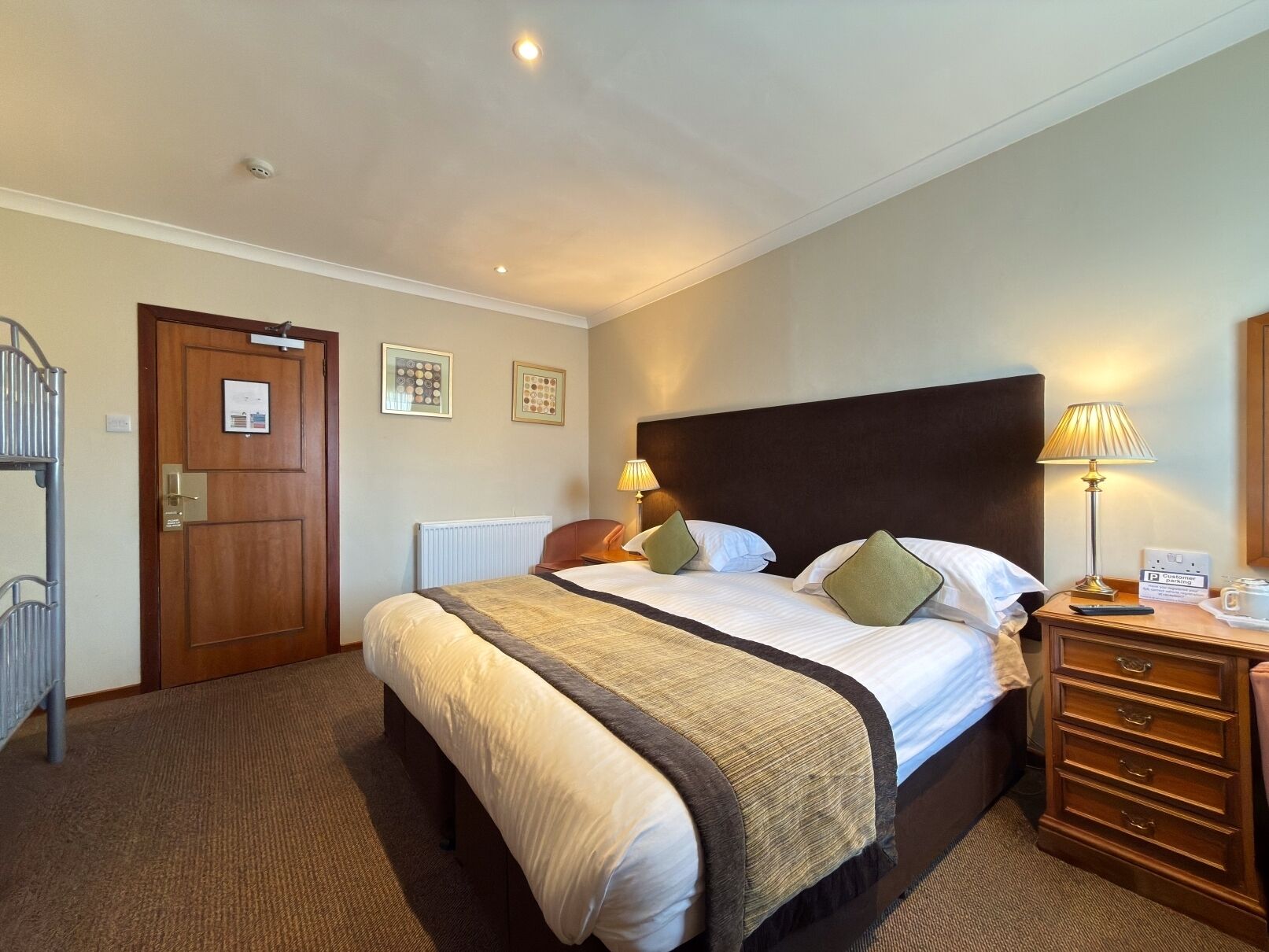 Habitación de hotel con cama, mesitas de noche, radiador, puerta y lámpara fijada a la pared.