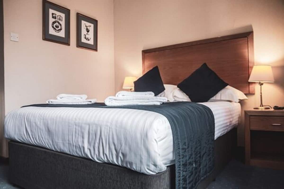 A neatly made bed with pillows and a folded towel, a wooden headboard, a bedside table with a lamp, and framed pictures on the wall.