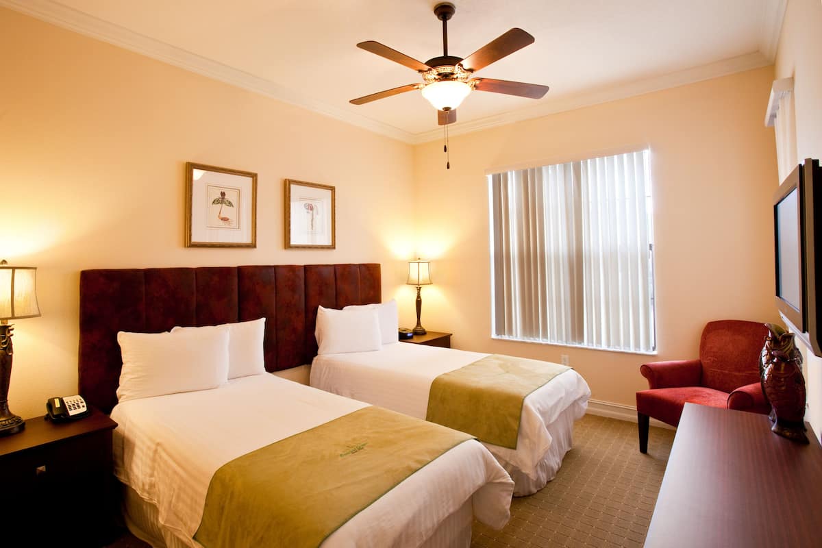 Een hotelkamer met twee bedden, een plafondventilator, een raam met zonwering en een rode stoel.