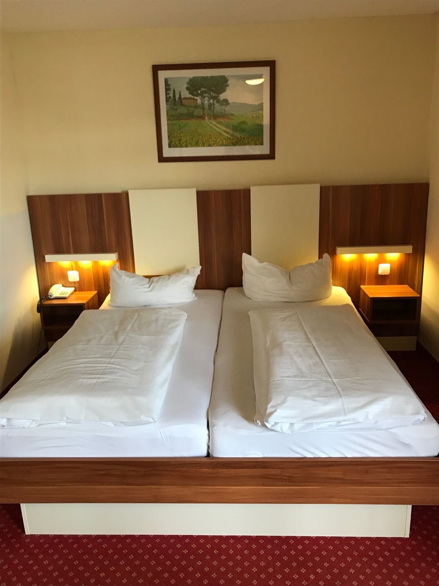 Un lit double avec du linge de lit blanc, une tête de lit en bois, deux tables de chevet avec des lampes et, au-dessus, un tableau représentant un paysage encadré.