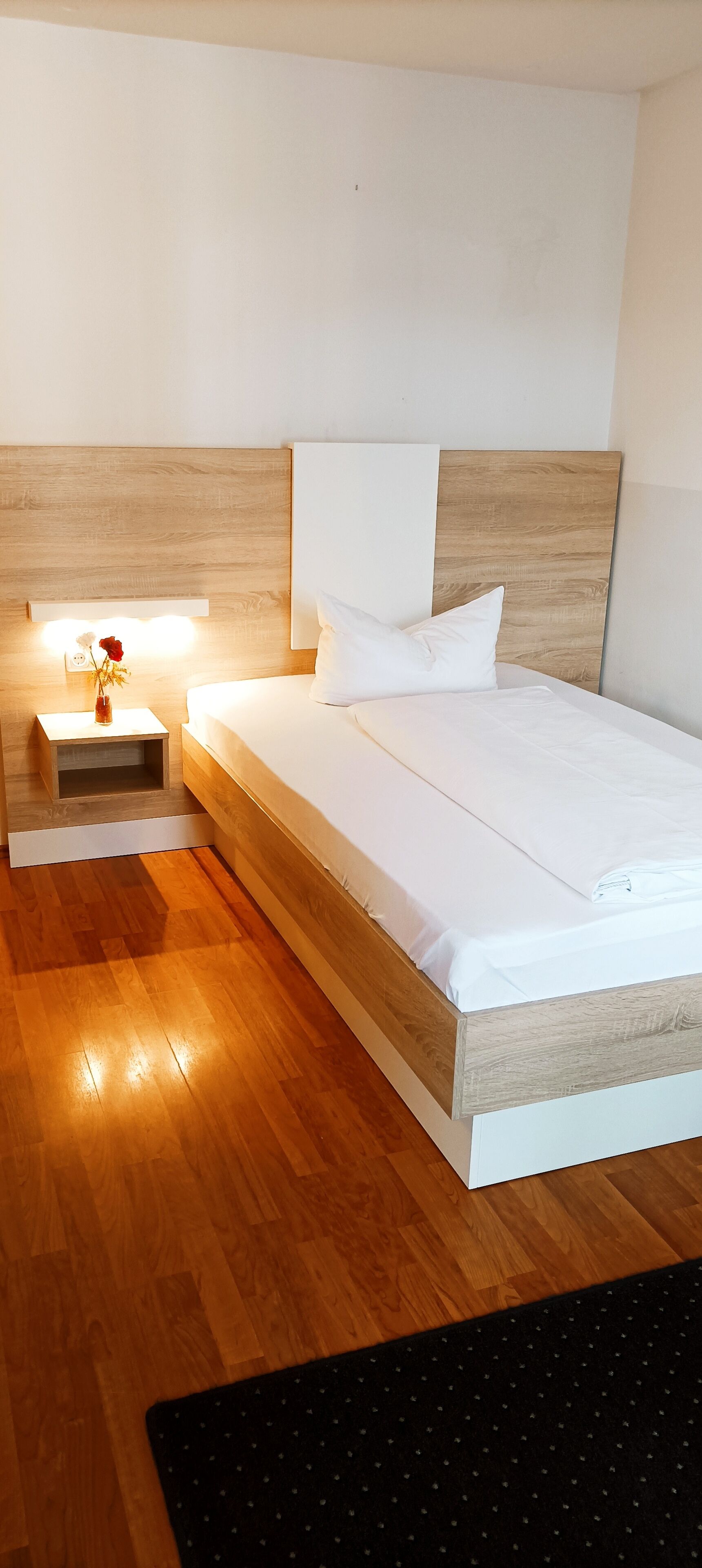 Une chambre d’hôtel moderne avec une tête de lit en bois, un lit parfaitement fait, une table de chevet ornée d’un vase de fleurs et un tapis noir sur le sol.