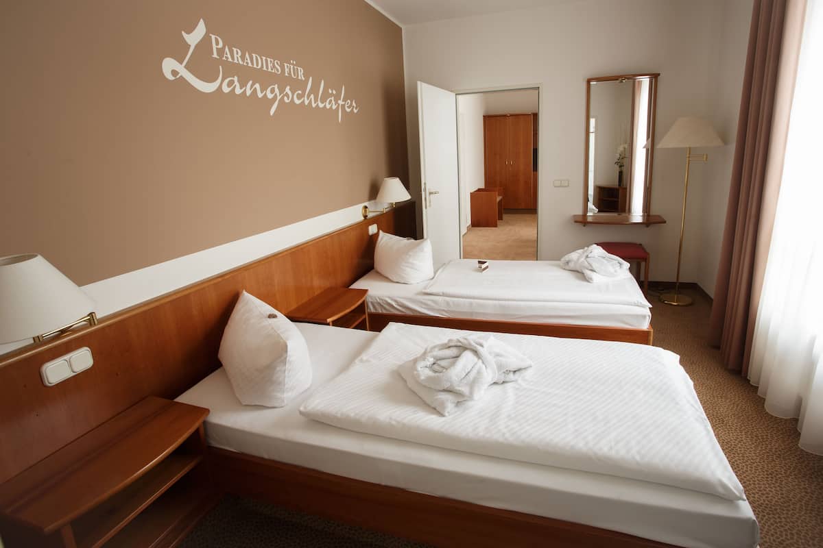 A hotel room with two beds, a wooden headboard, a bedside table with a lamp, a mirror, and a carpeted floor.