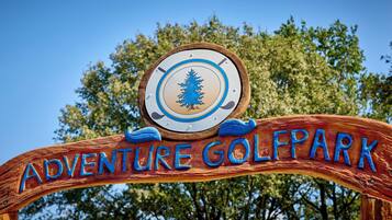 Ein hölzernes Schild mit der Aufschrift 'ADVENTURE GOLF PARK' und einem kreisförmigen Emblem mit einem Baum.