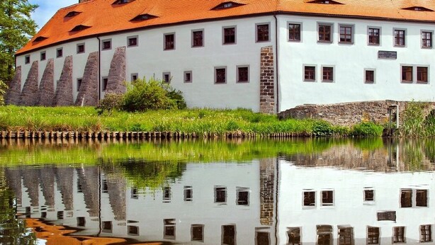 A large, white building with a red roof, reflecting in a calm body of water. - Hotel Sportwelt (Radeberg)