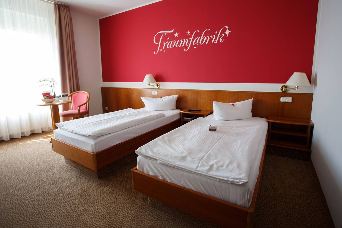 A hotel room with two beds, a red wall with a 'Traumfabrik' sign, a wooden headboard, a small table with a lamp, and a chair.