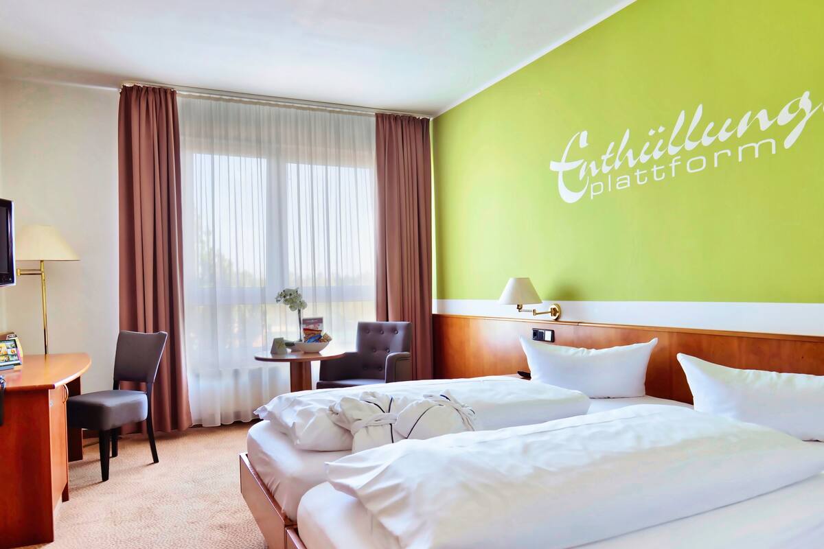 A hotel room with two beds, a desk, a chair, a small table with a lamp, and a television.