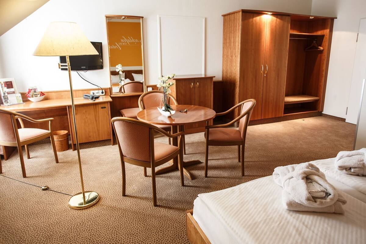 A hotel room with a bed, a desk with a lamp, a chair, a table, a wardrobe, and a mirror.