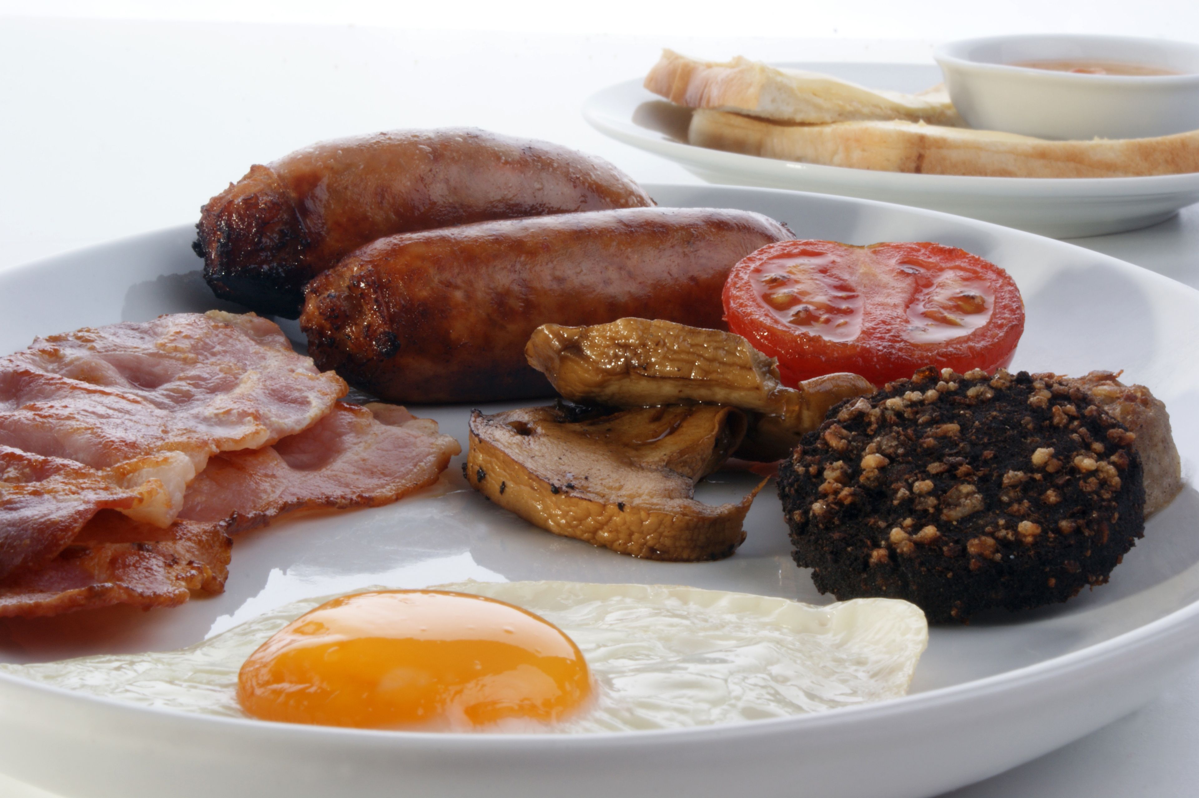 A plate of breakfast food including sausages, bacon, mushrooms, a tomato slice, a fried egg, and black pudding.