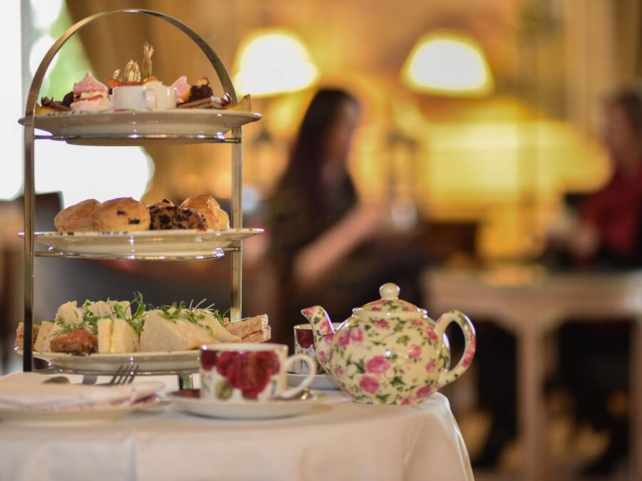 A three-tiered tea service stand with various pastries and sandwiches, a floral teapot, and a cup on a table in a warmly lit room.