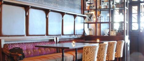 A restaurant with a long bench seating area, a bar with stools, and a display case with glassware.