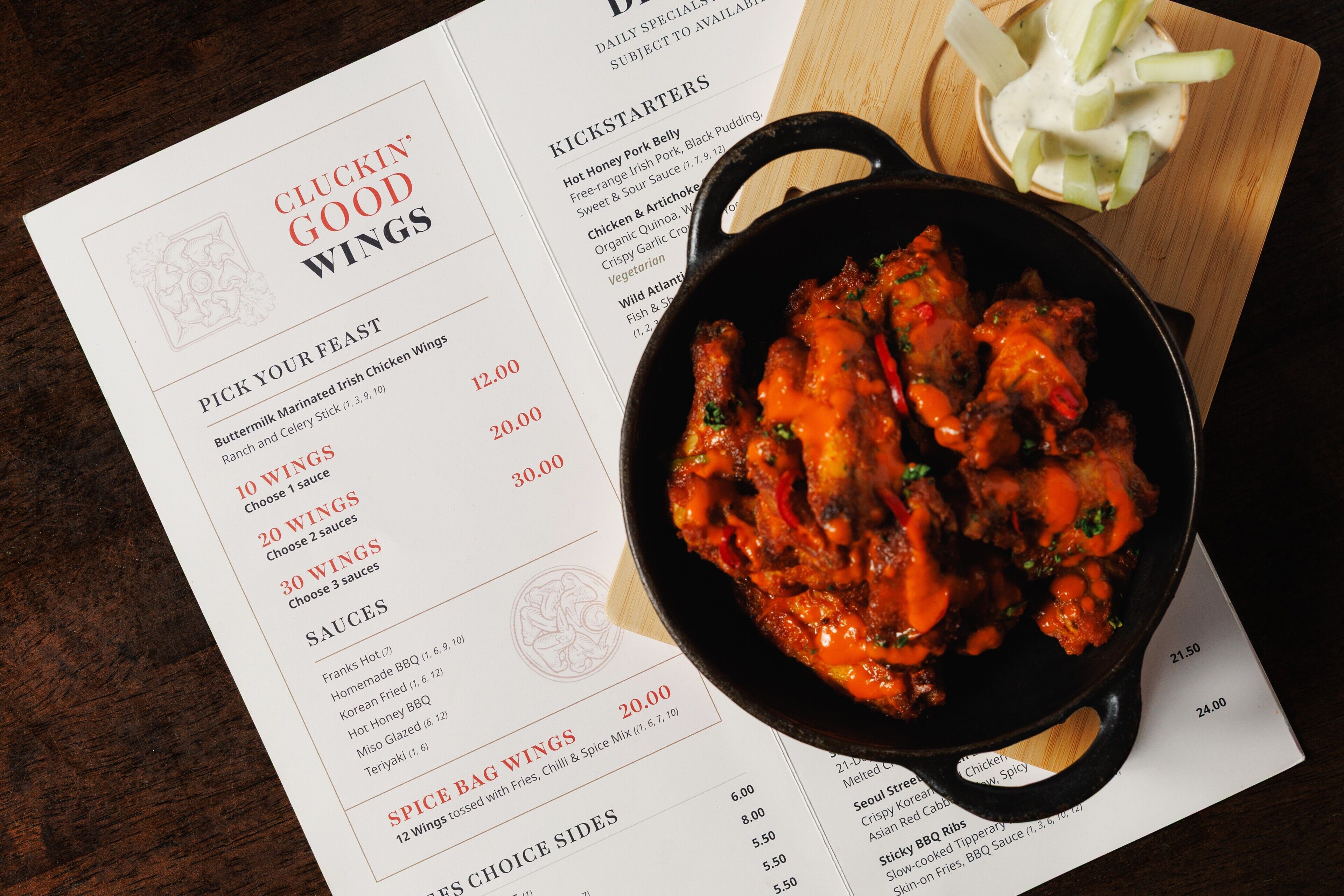 Eine Speisekarte mit verschiedenen Gerichten und Preisen, eine Schüssel mit Chicken Wings und eine Holzplatte mit Selleriestangen.