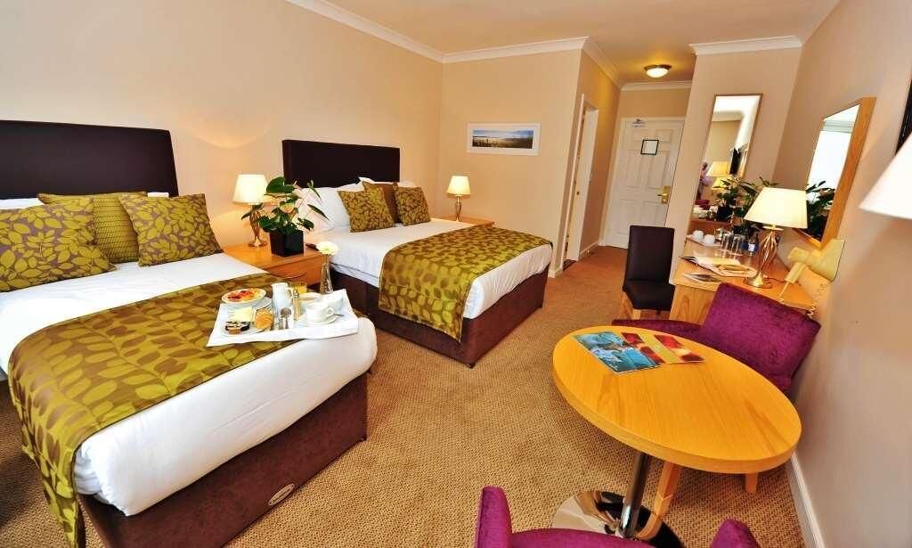 A hotel room with two beds, a small round table, a chair, and a desk with a lamp.