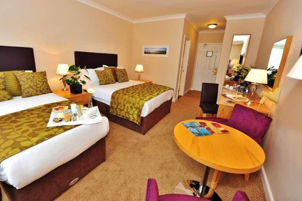 A hotel room with two beds, a small round table, a chair, and a desk with a lamp.