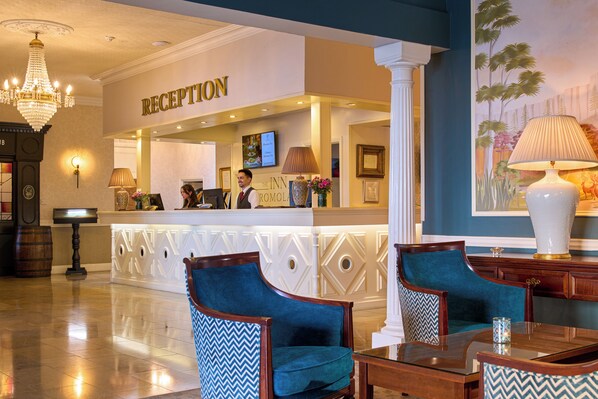 A reception area with a reception desk, two blue upholstered chairs, a table, and a chandelier.
