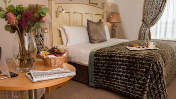 Habitación de hotel con una cama bien hecha, una mesita de noche con un periódico, una cesta de frutas y un florero con flores.