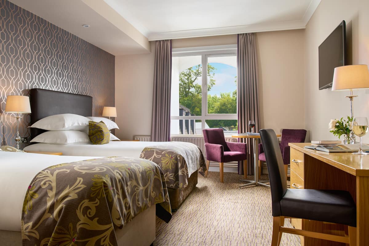 Une chambre d’hôtel avec deux lits, un bureau, une chaise, une fenêtre avec des rideaux et une vue sur la verdure.