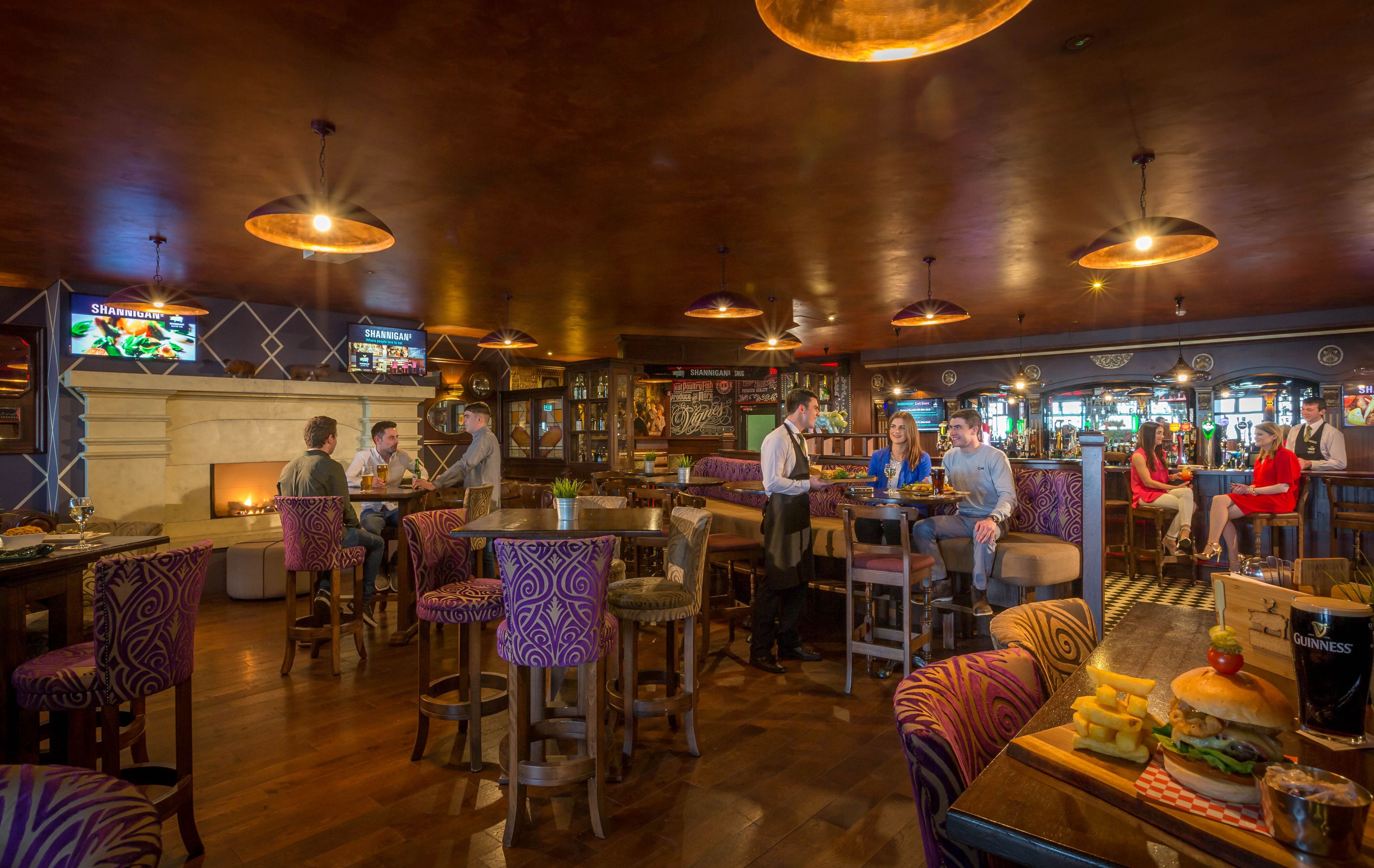 Un bar avec des meubles en bois, des luminaires suspendus et un tableau affichant le menu.