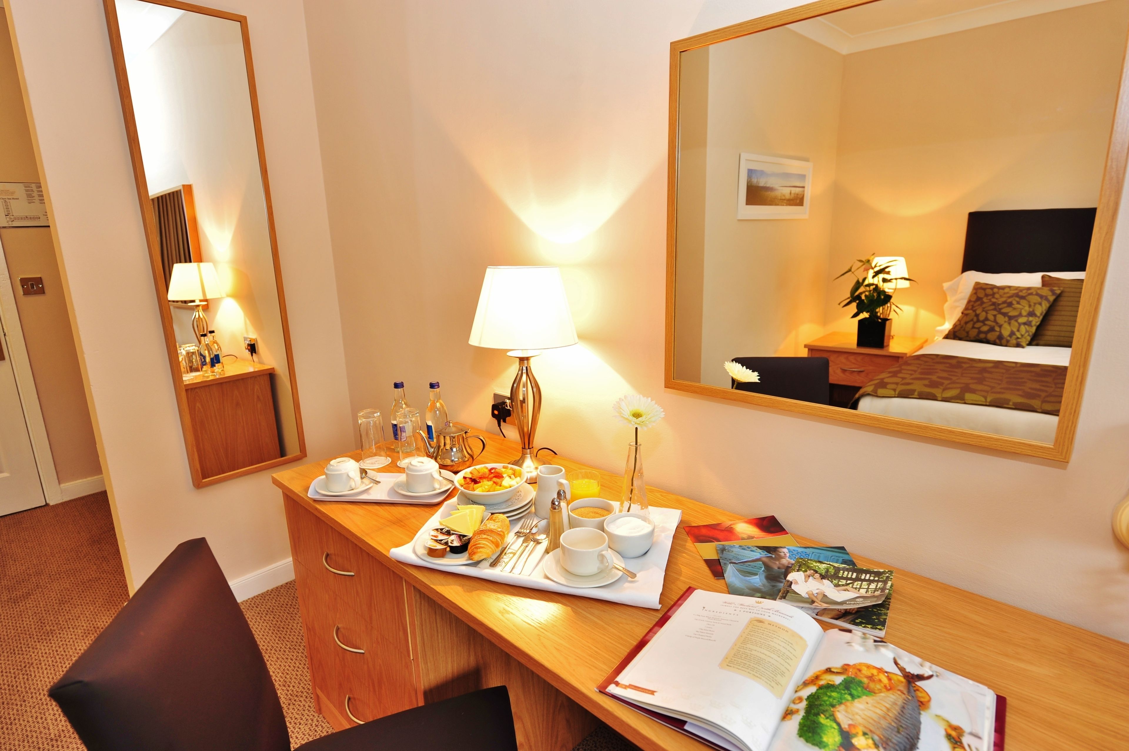 A hotel room with a wooden desk, a mirror, a bed, and a bedside table with a lamp and a plant.