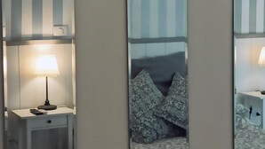 A room with a bed, a bedside table with a lamp, and a mirror reflecting the bed and a nightstand with a lamp. - Dei Decumani (Naples)