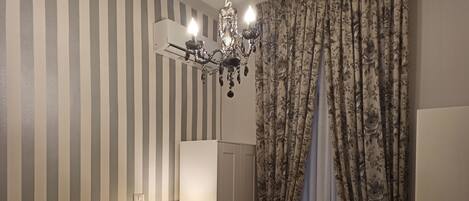 Une chambre d’hôtel avec un mur à rayures, un lustre, un lit avec une literie à motifs, une table de chevet avec une lampe et des rideaux à fleurs.