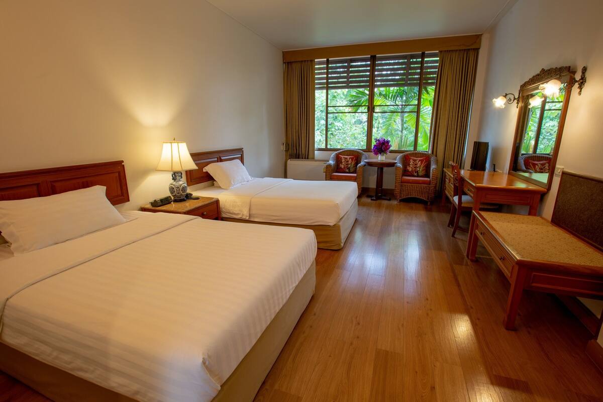 A hotel room with two beds, a desk, a chair, a small table with a lamp, and a window with curtains.