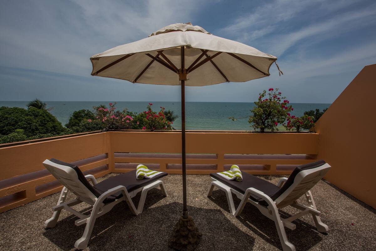 A balcony with two lounge chairs and a large umbrella, overlooking the sea.