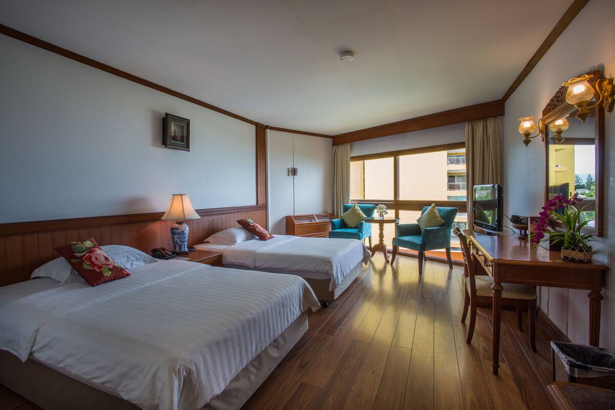 A hotel room with two beds, a desk, a chair, a lamp, and a window with a view of the outside.