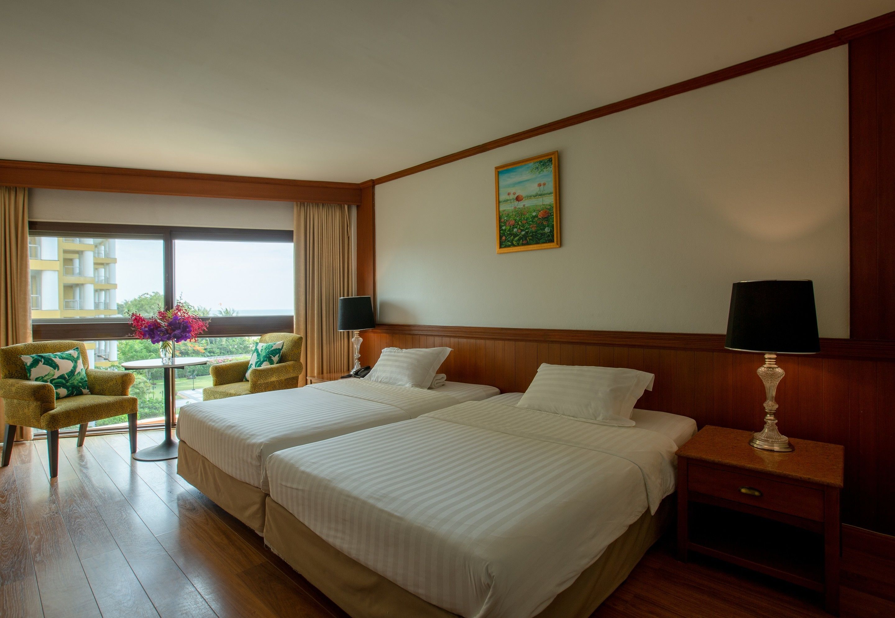 A hotel room with two beds, a wooden floor, a window with curtains, a small table with a vase of flowers, and a painting on the wall.