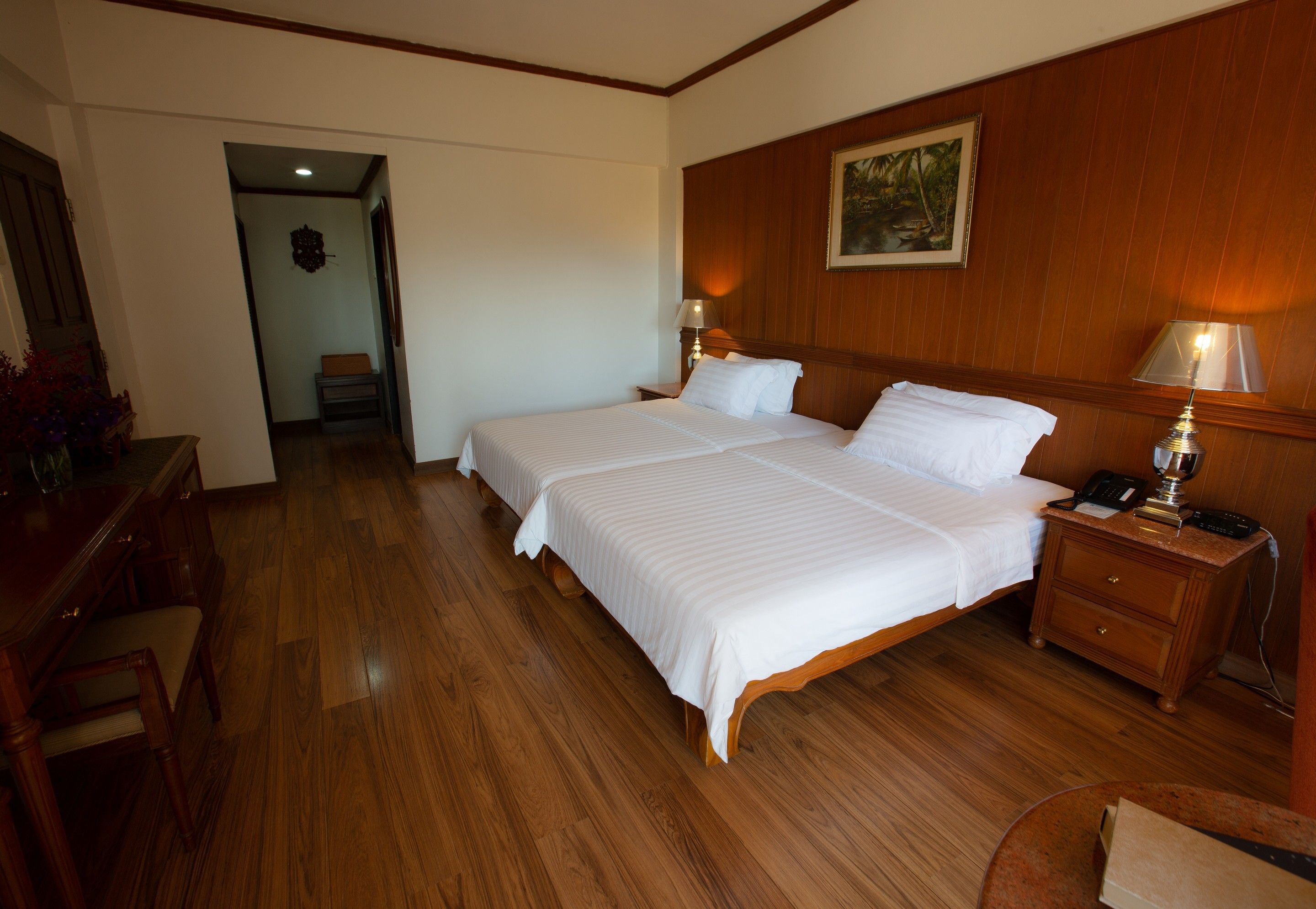 A hotel room with two beds, wooden flooring, a desk with a chair, a nightstand with a lamp, and a painting on the wall.