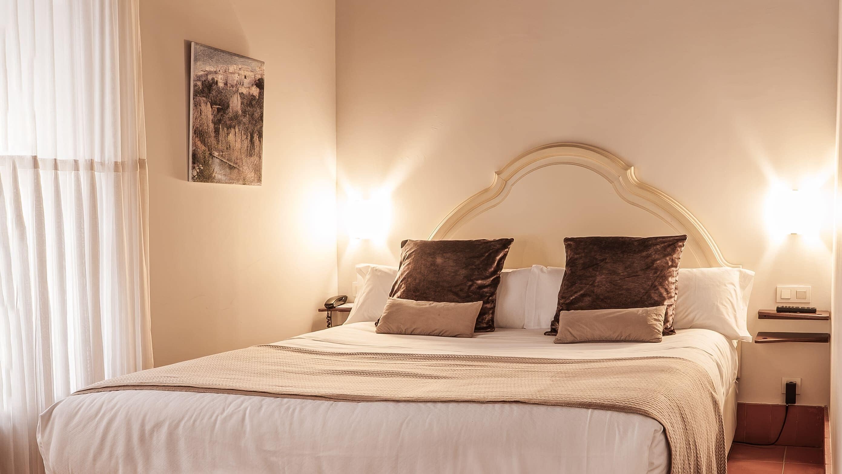 Una cama bien hecha con cabecera, dos mesitas de noche, un cuadro colgado en la pared y una ventana con cortinas translúcidas.