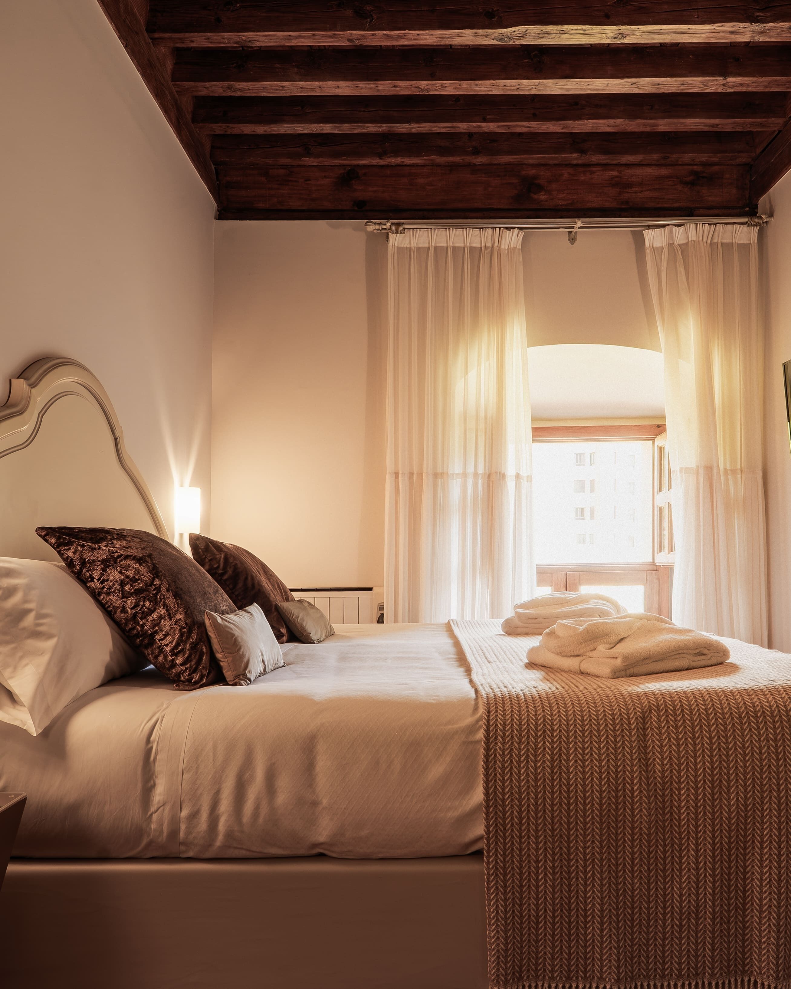 Un dormitorio con techo de madera, una cama con ropa de cama blanca, una mesita de noche y una ventana con cortinas de gasa.