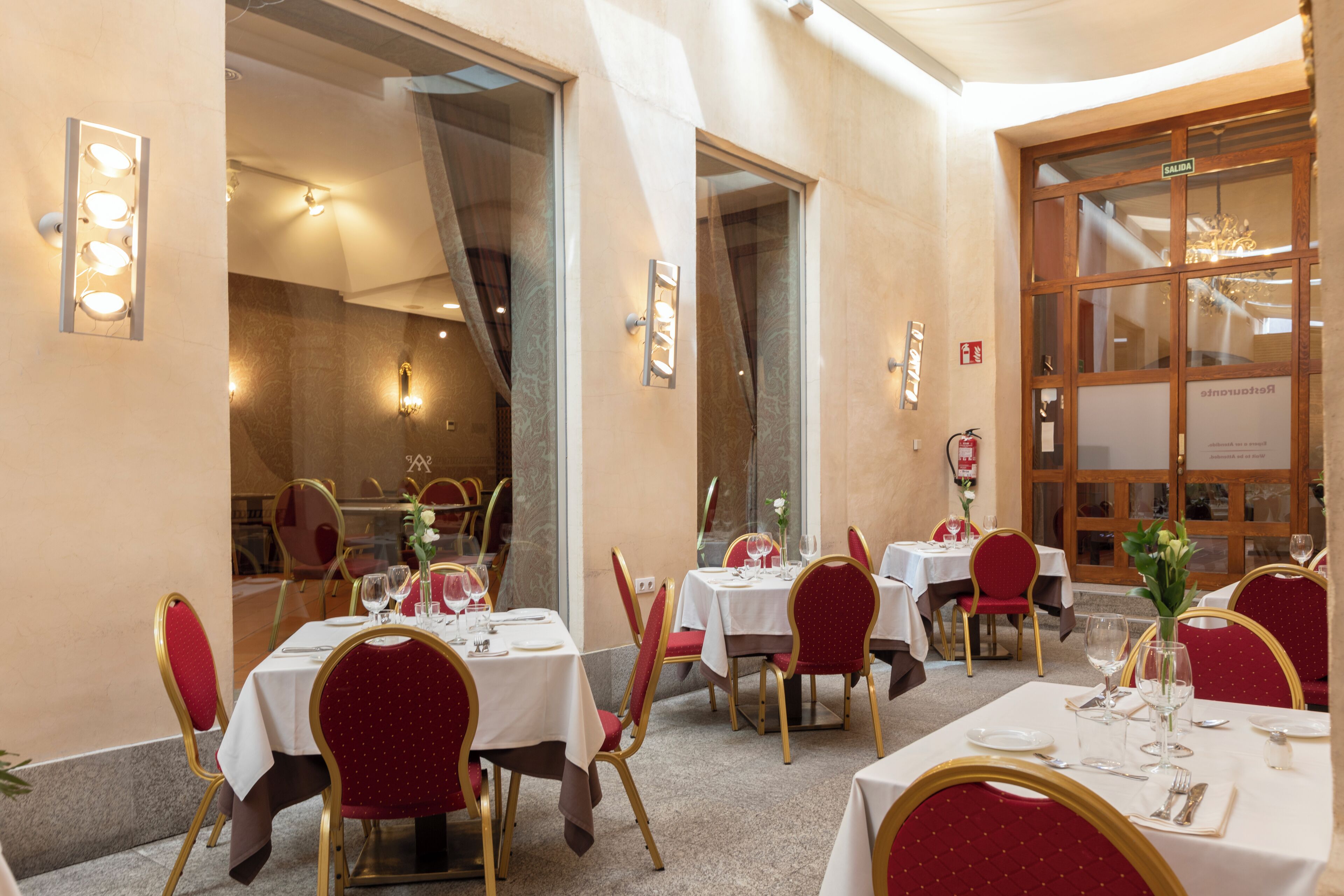 Un comedor con mesas dispuestas para el servicio, sillas tapizadas en rojo y una araña visible a través de un ventanal amplio.