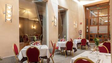 Une salle à manger avec des tables dressées, des chaises à l’assise rouge et un lustre visible à travers une grande fenêtre.