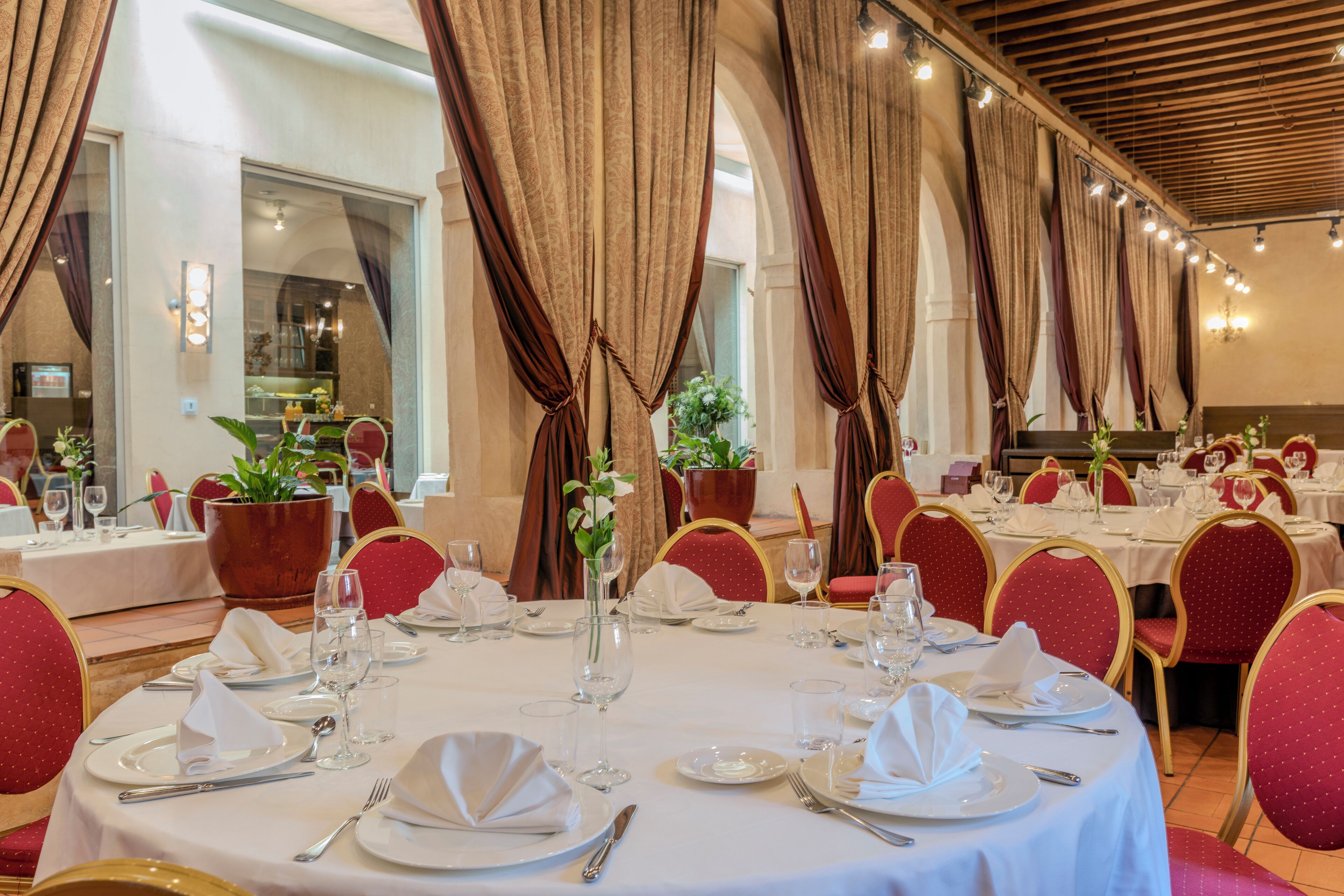 Comedor con mesas redondas dispuestas para una comida, sillas tapizadas en rojo y ventanas amplias con cortinas.