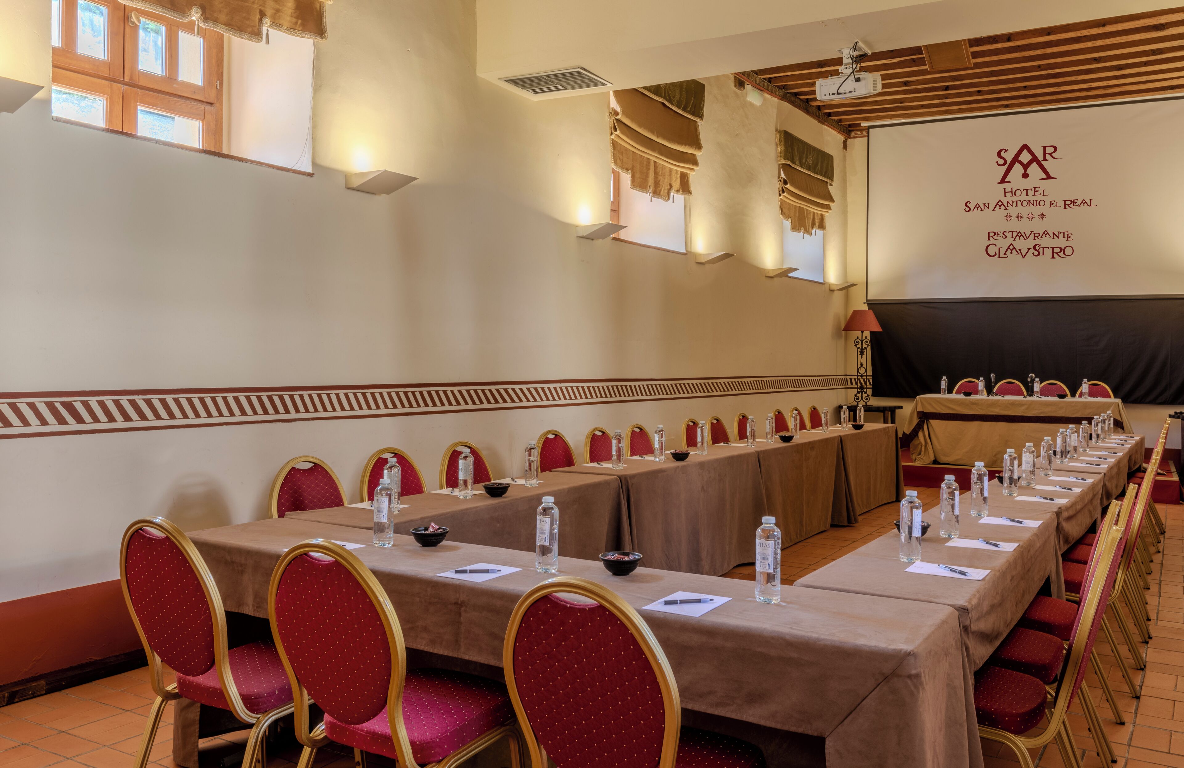 A conference room with a long table, red chairs, and a projector screen displaying the hotel's name.
